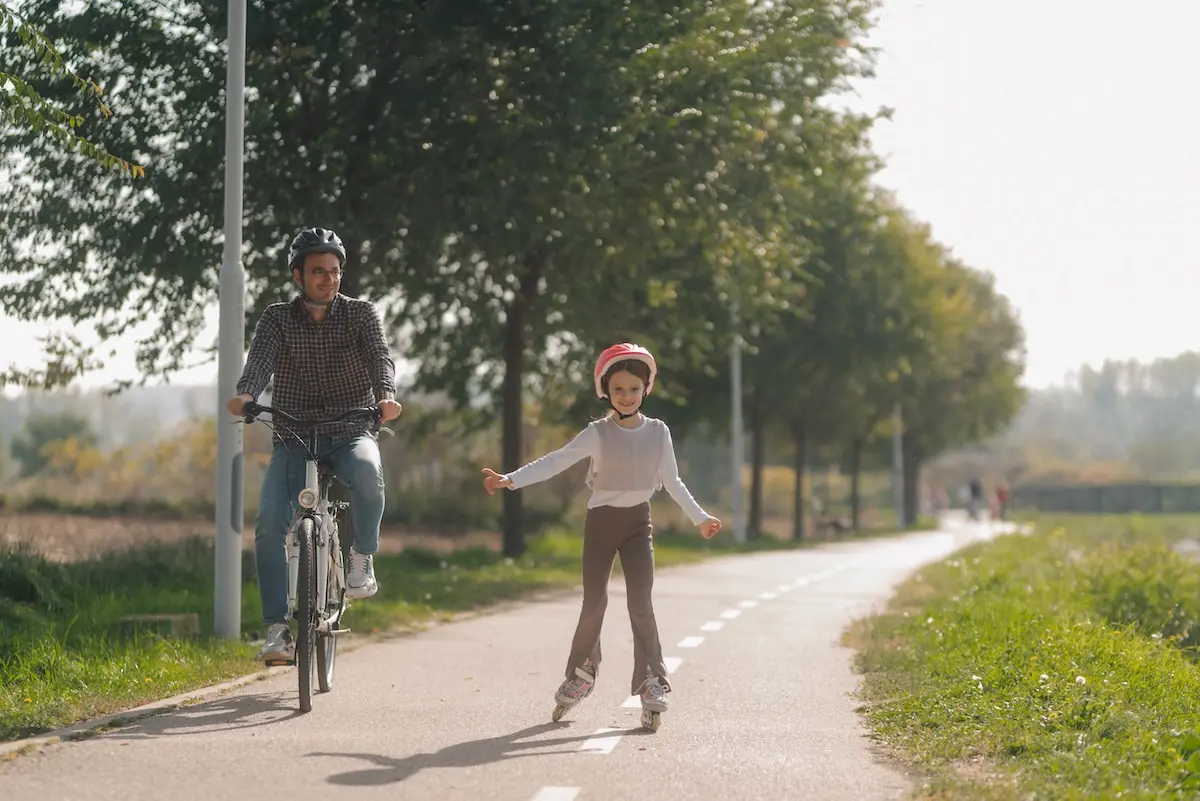 A dad on a bike next to a girl on rollerblades, traveling on a bike path