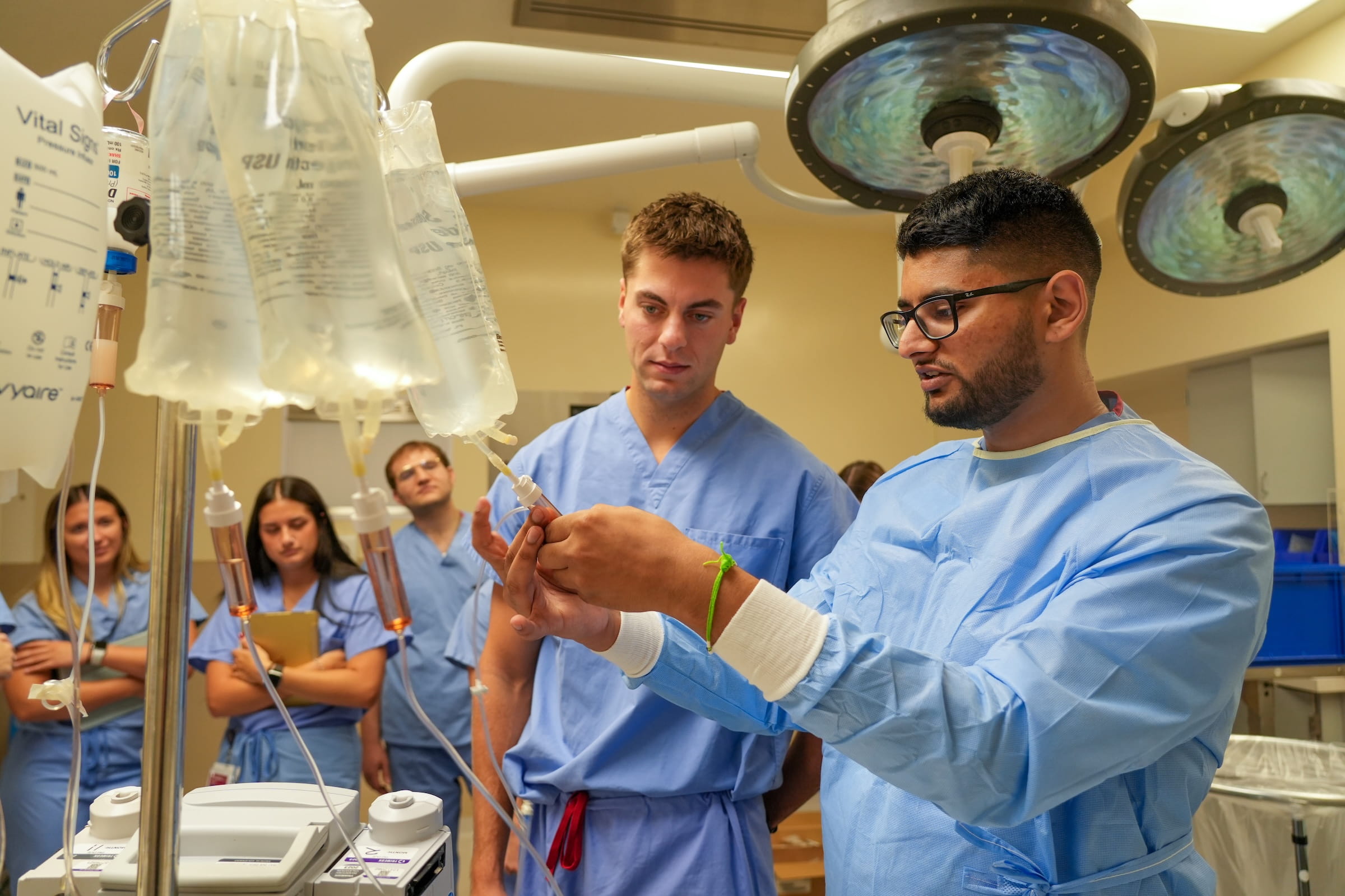 Students wearing scrubs work with IV's on poles in a simulated operating room