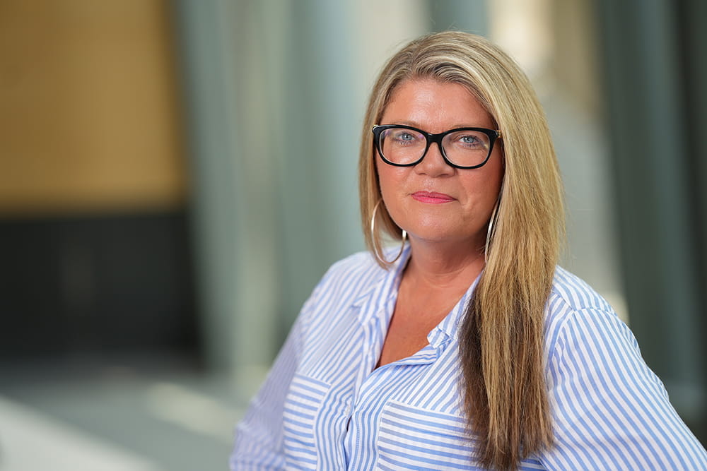 Head and shoulders shot of Courtney Cowley, woman with blonde hair wearing dark glasses and blue striped blouse