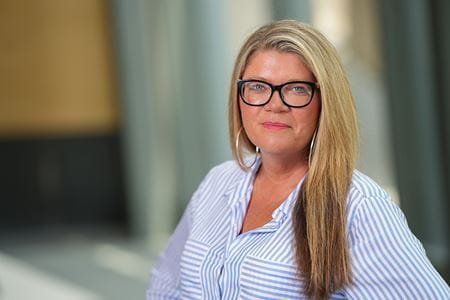 Head and shoulders shot of Courtney Cowley, woman with blonde hair wearing dark glasses and blue striped blouse