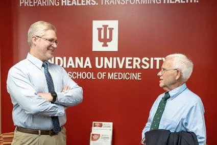 Dr. Matthew Tews stands with arms crossed, smiling and talking with Dr. Patrick O'Neil in front of a red wall with the IU School of Medicine logo on the West Lafayette campus.