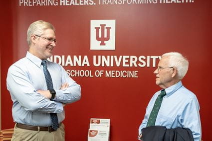 Dr. Matthew Tews stands with arms crossed, smiling and talking with Dr. Patrick O'Neil in front of a red wall with the IU School of Medicine logo on the West Lafayette campus.