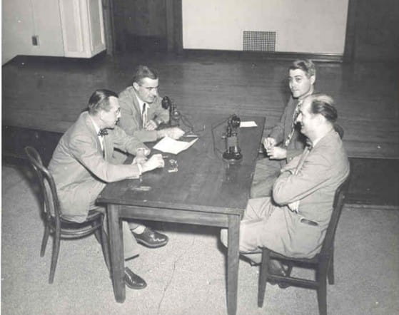 Title: first telephone transmitted class in 1950; Four men sitting at a table, speaking into a telephone to teach class