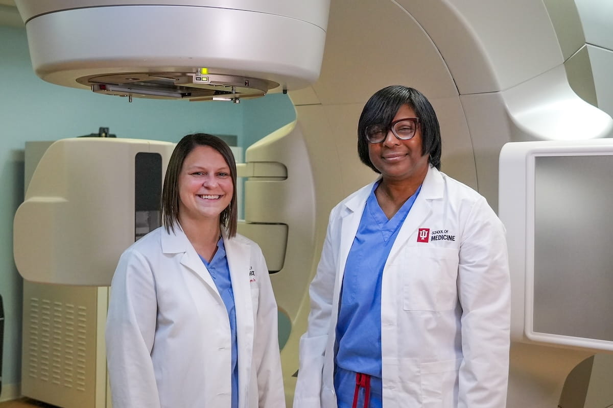 Megan Knight and Maria Walker, in white coats, stand in front of a radiation therapy machine in a treatment room.