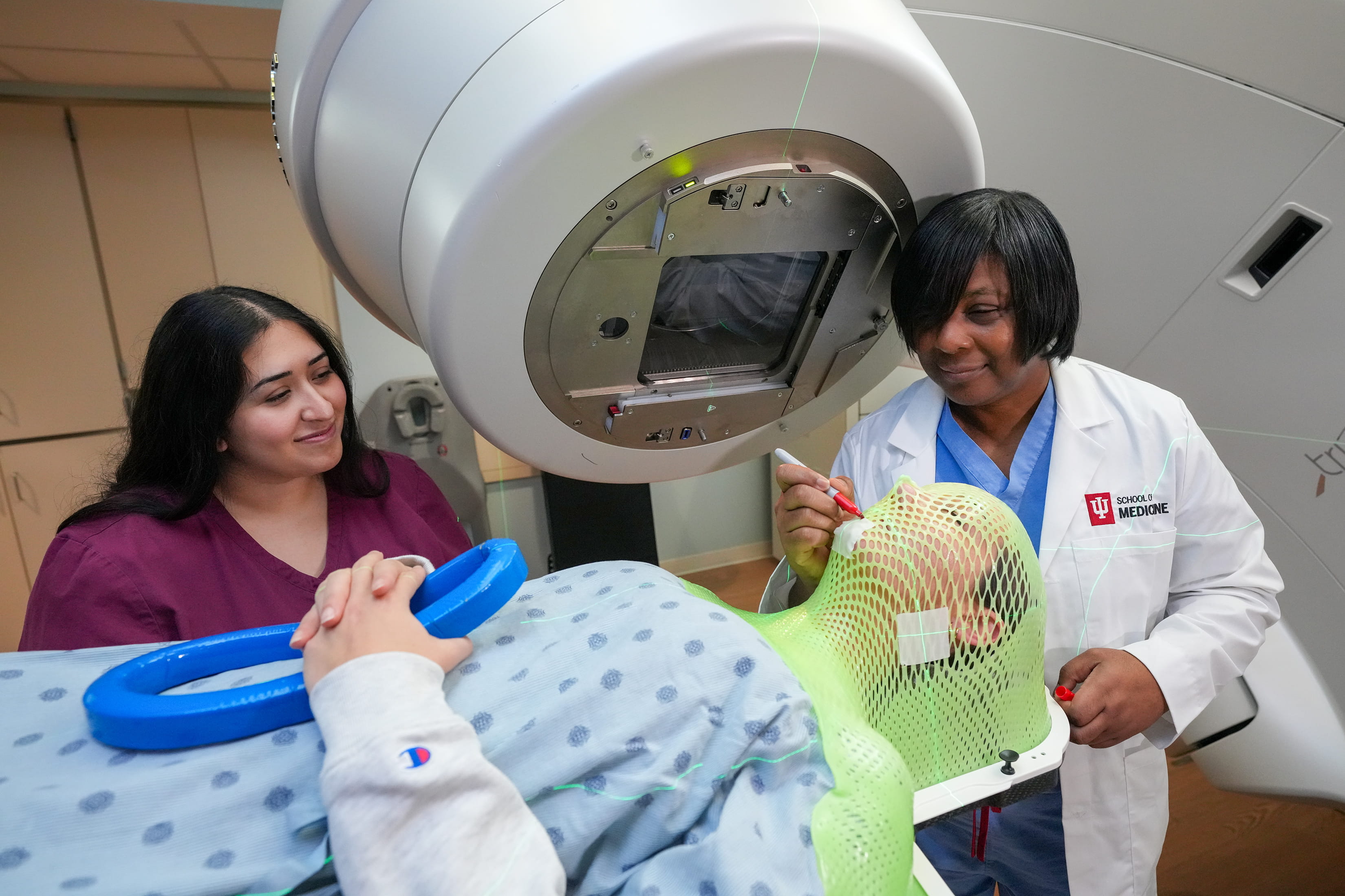 A student in hospital gown lies on a table with a green thermoplastic radiation therapy mask on as program director Maria Walker marks spots for mock treatment and another student looks on.