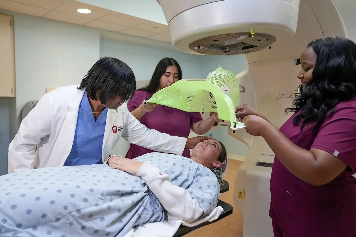 Maria Walker, in white coat, with three students: one lying on the table and two placing a radiation therapy mask.