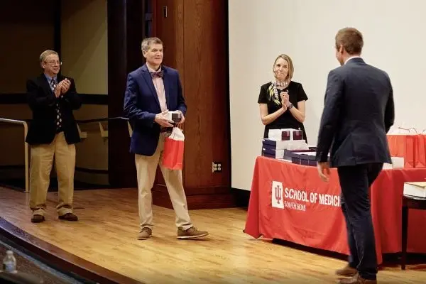 Joseph Harmon about to present a red bag to a graduate student as other school leadership applaud. 