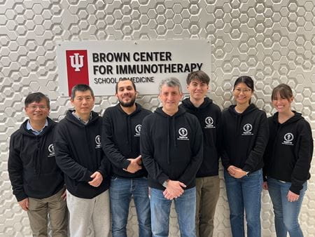 Mark Kaplan with six members of his lab wearing black Kaplan Lab sweatshirts in front of the Brown Center for Immunotherapy sign