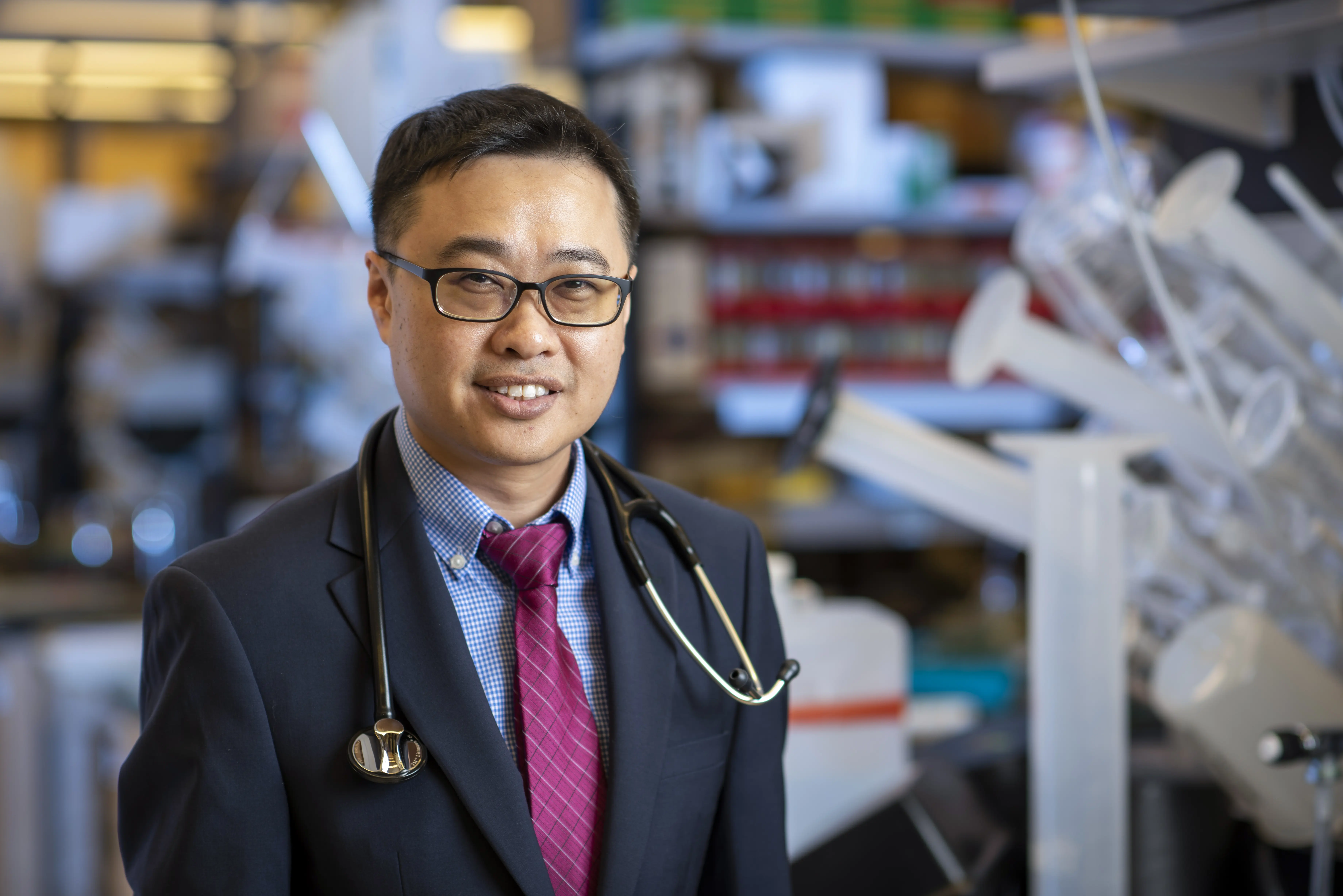A man with short dark hair and glasses smiling in a laboratory setting while wearing a suit with a stethoscope around his neck.