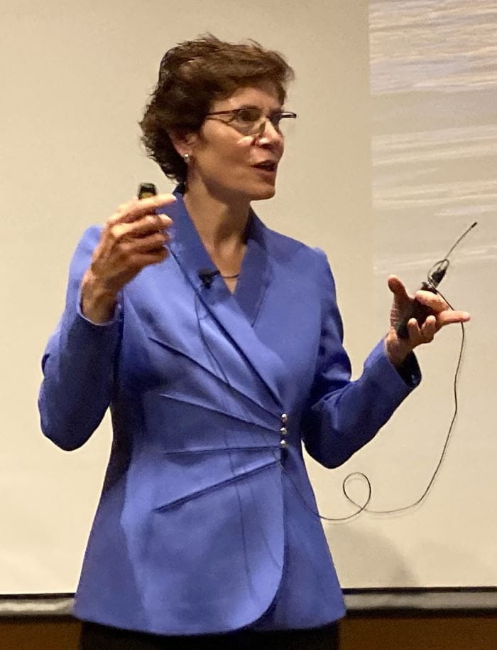 Woman speaks in front of an audience, holding a portable mic.