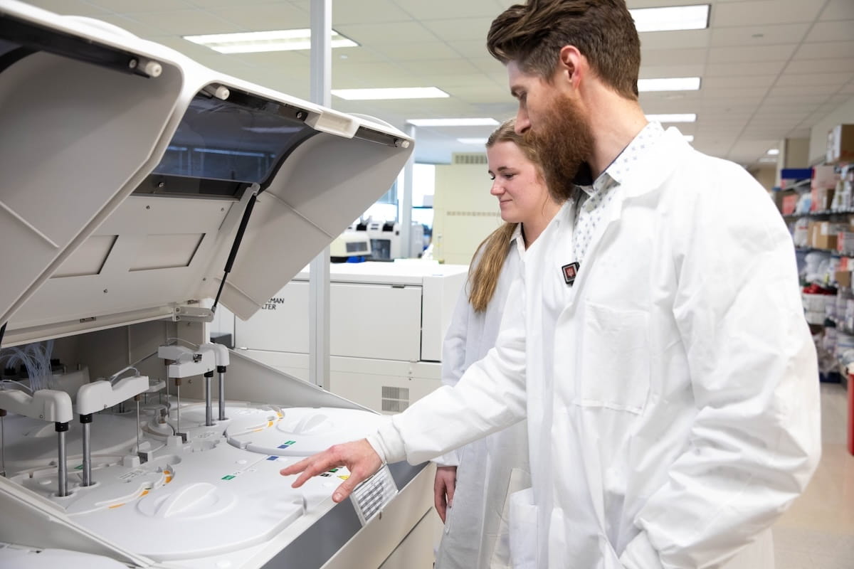 Nicholas Brehl trains a student, both wearing lab coats, on equipment in the laboratory.