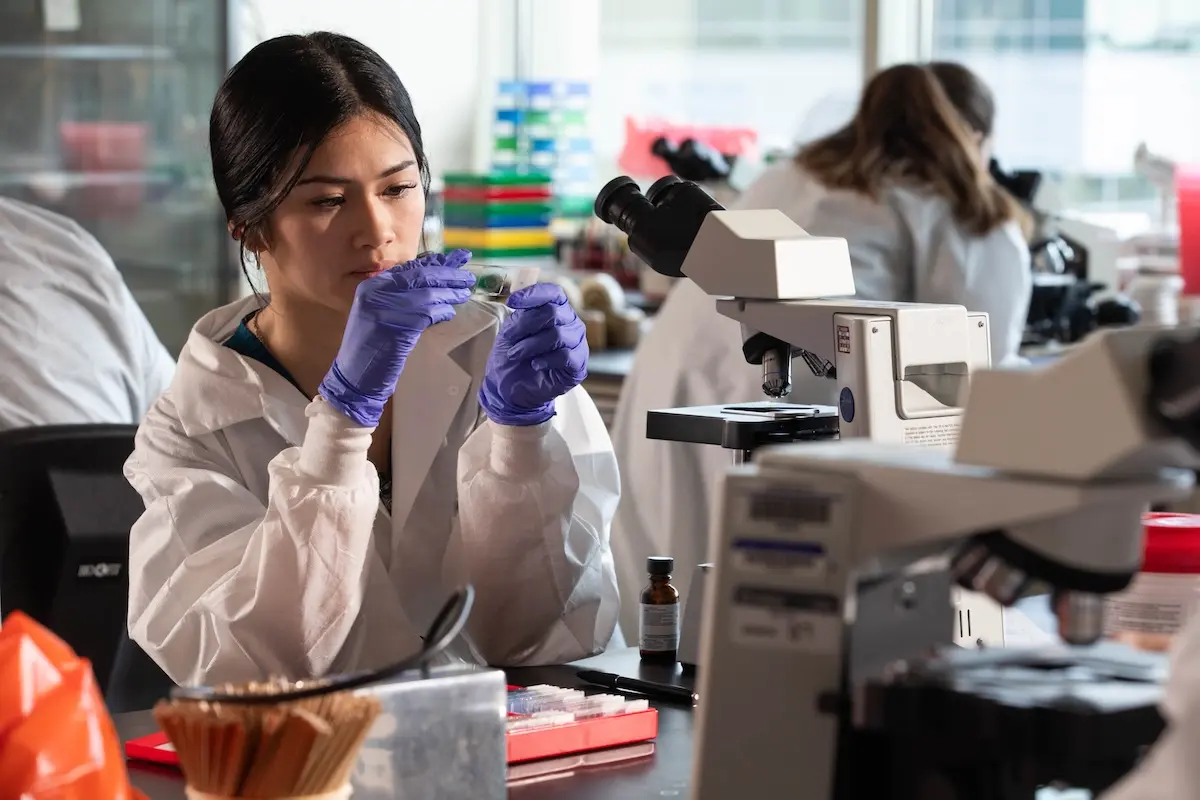 Female student in lab coat and gloves examines a specimen near a microscope