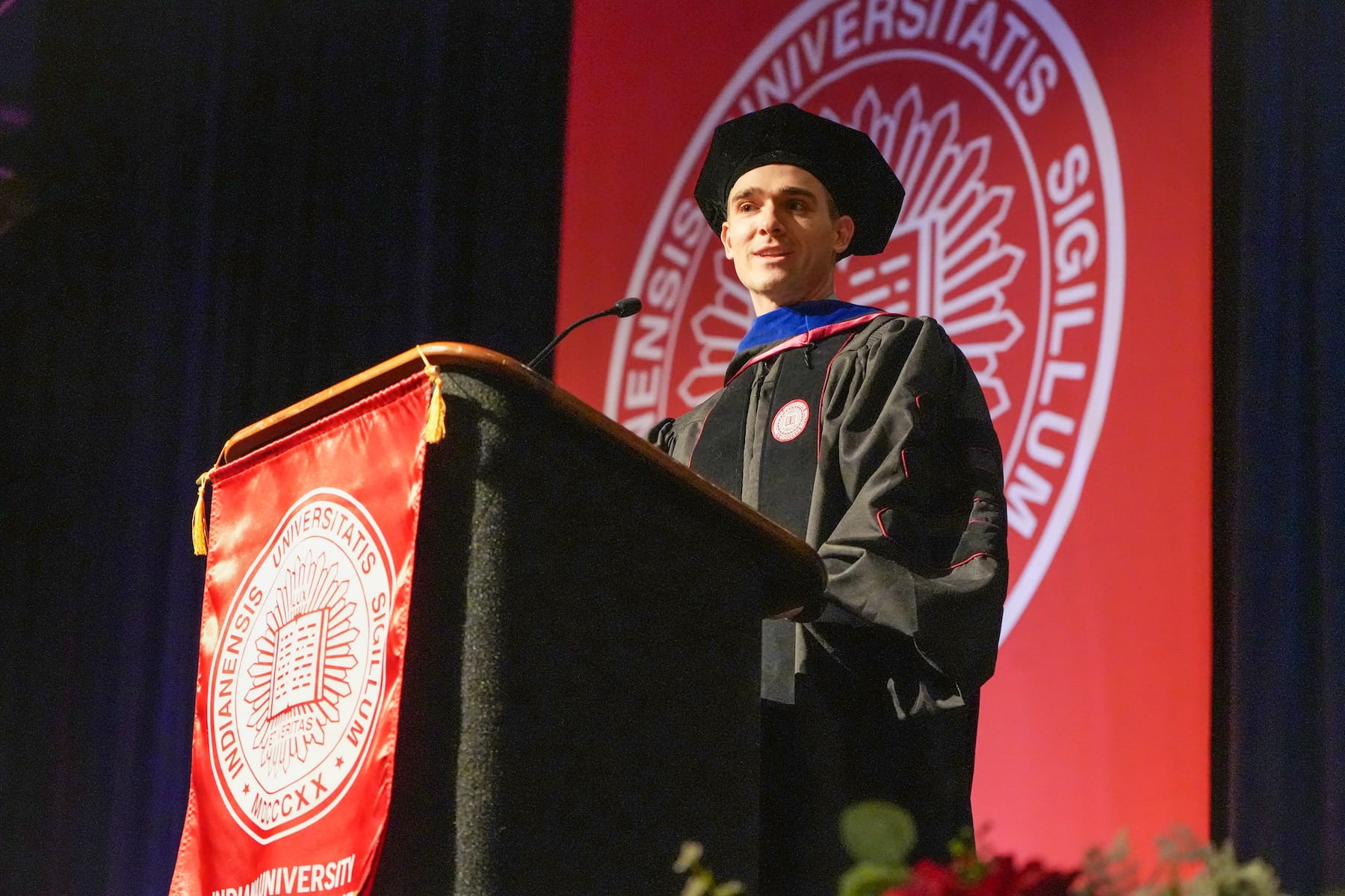 Oliver Strobel speaks from the podium at graduation, wearing his graduation regalia
