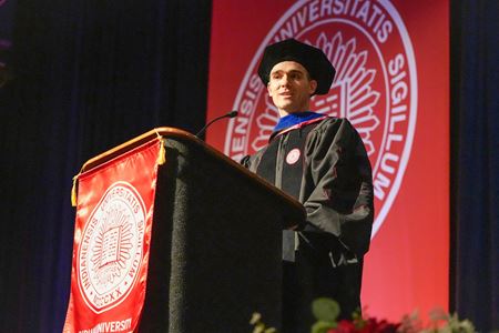Oliver Strobel speaks from the podium at graduation, wearing his graduation regalia