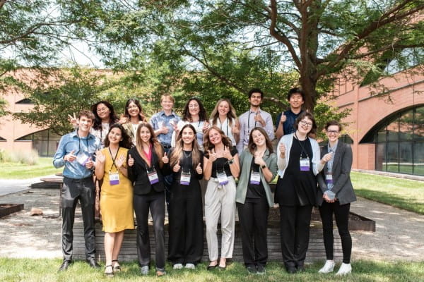 about 15 students with conference nametags around their necks stand outside on a summer day and give thumbs ups