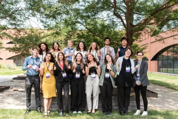 about 15 students with conference nametags around their necks stand outside on a summer day and give thumbs ups