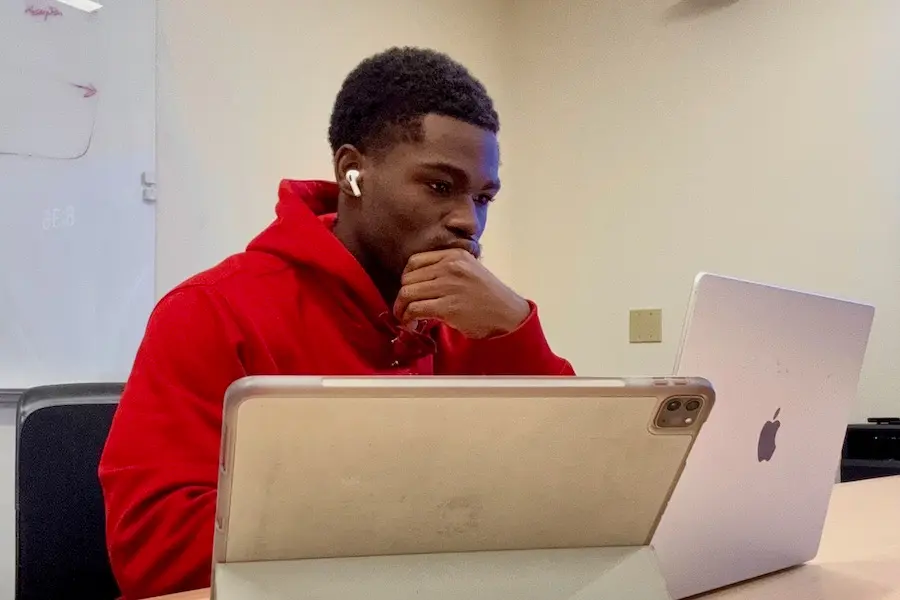 Mout-Maine Moustapha, Black man in red hoodie, sits at his computer, wearing ear pods.