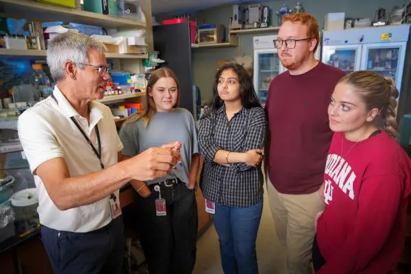 John Turchi holding an object in the lab while speaking to a group of four lab workers.