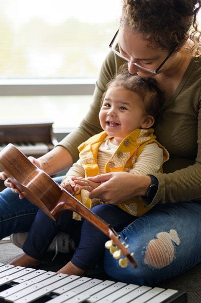 a smiling young child sits in her mother's lap holding a ukelele