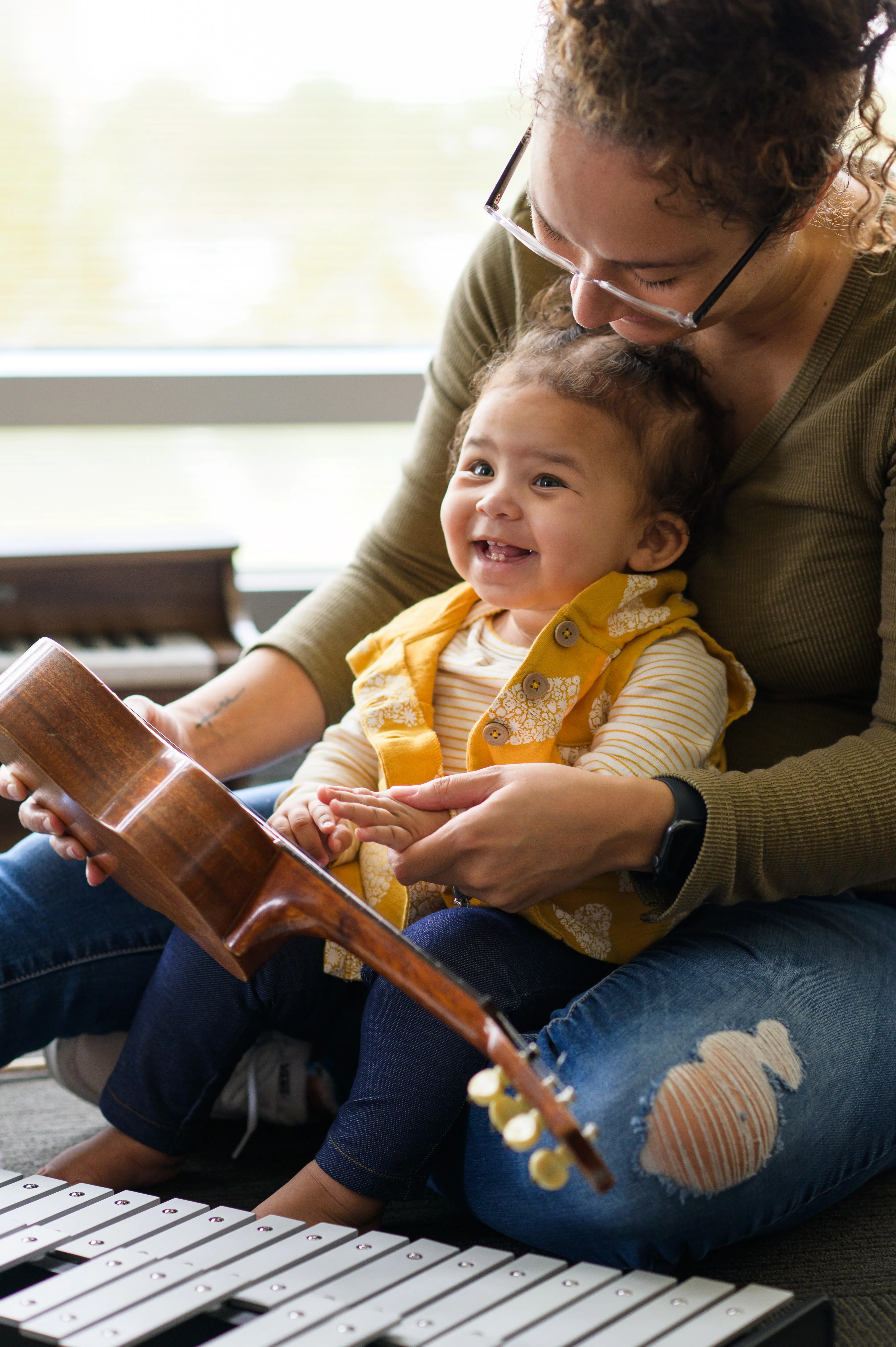 a smiling young child sits in her mother's lap holding a ukelele