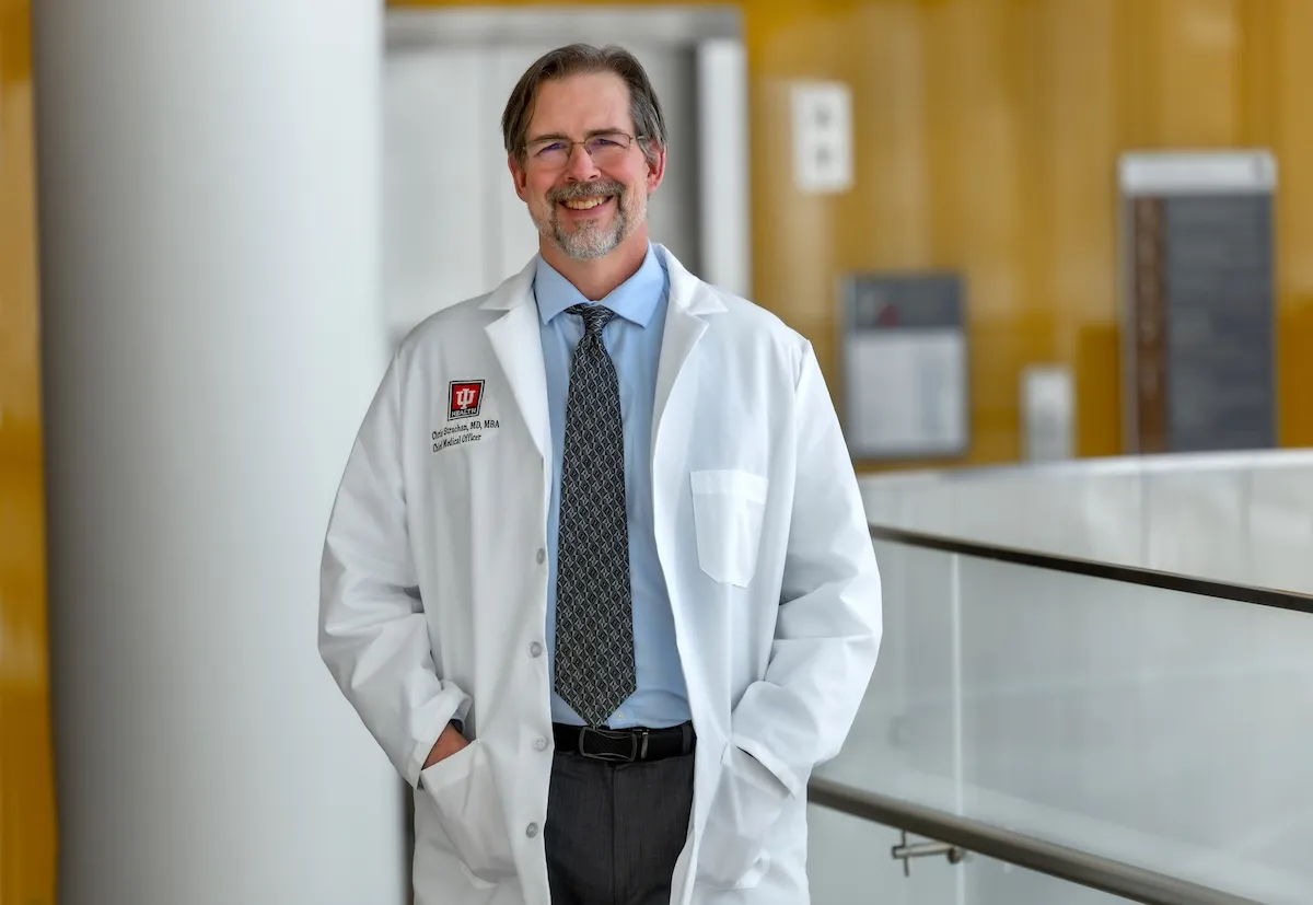 Dr Christian Strachan stands near a desk at an IU Health hospital wearing his white coat, a blue shirt and tie, with hands in his coat pockets