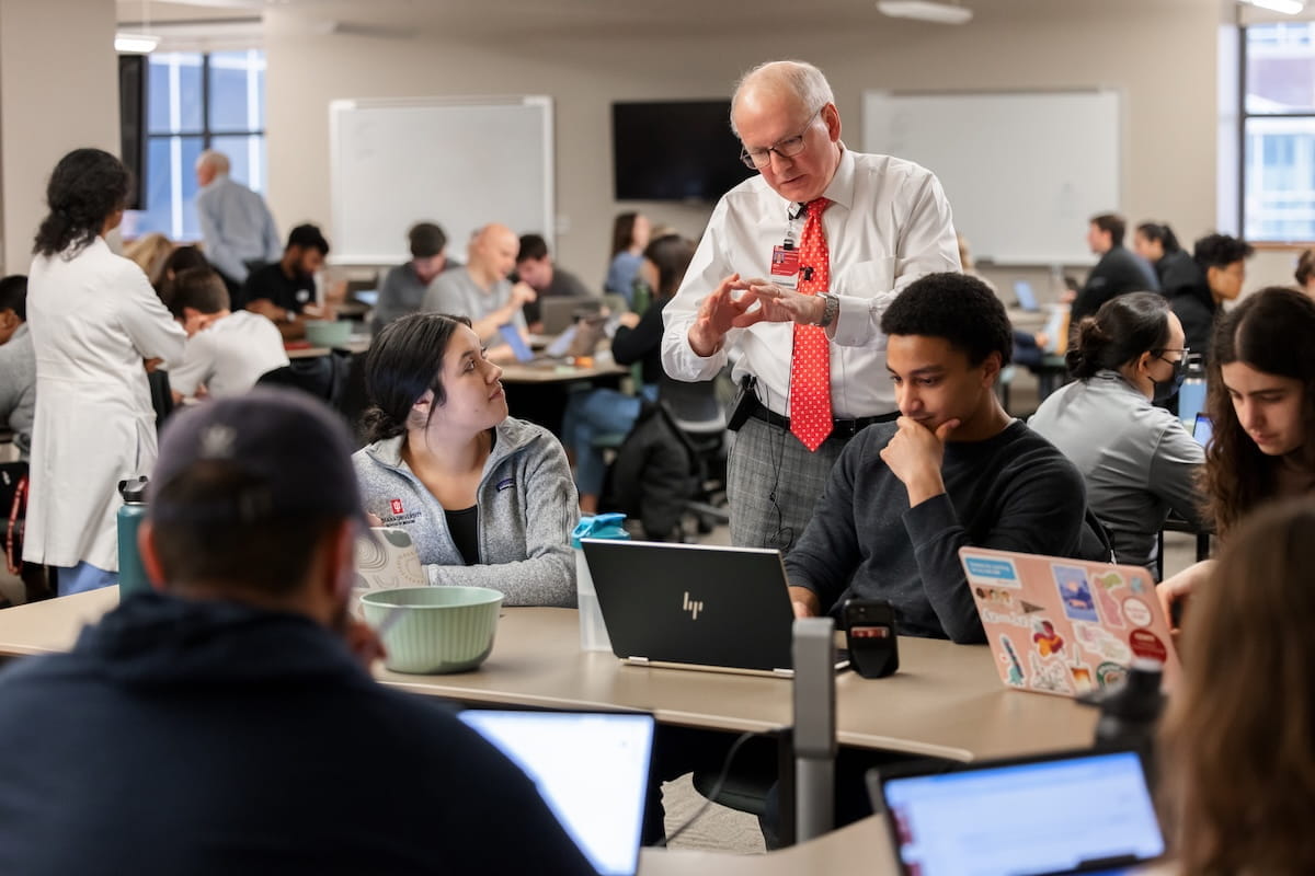 Dean Jay Hess stands beside a table of students working on laptops and instructs them