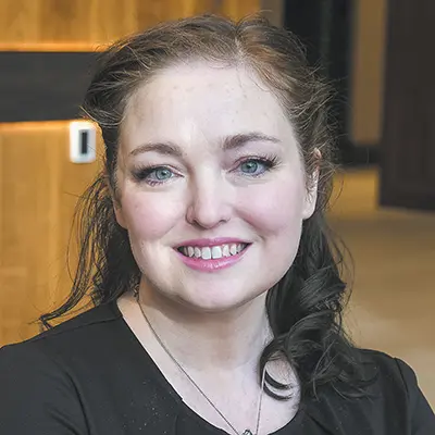 Headshot of a woman with wavy auburn hair worn loose, smiling, wearing a black top, photographed indoors against a wood-paneled wall. Photo/Image courtesy: Gabrielle Geddes