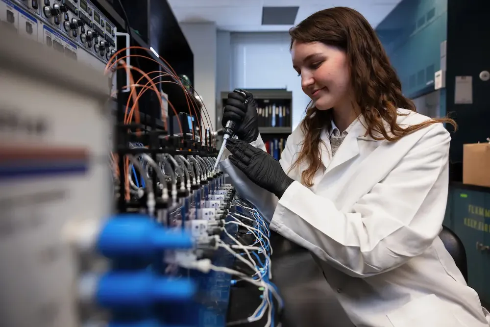 A female PhD student, wearing a white lab coat and black gloves, handles tubing on a machine in the IU Hydrocephalus Research Center.