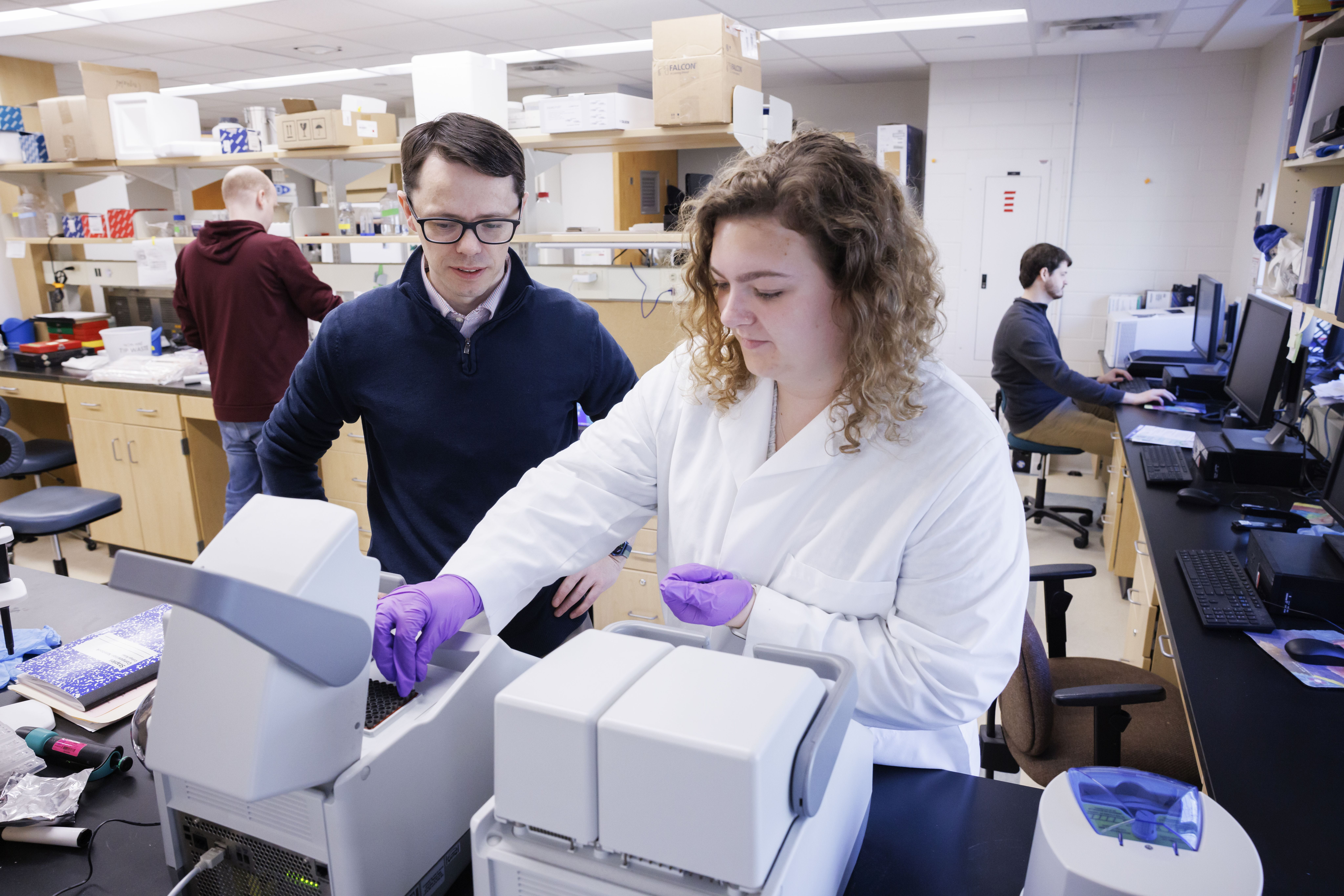 A laboratory scene where two individuals work together at a benchtop instrument, one wearing a lab coat and gloves while adjusting samples. Another individual observes closely. Additional researchers work in the background at lab benches and a computer workstation. Shelving above holds containers and lab supplies.