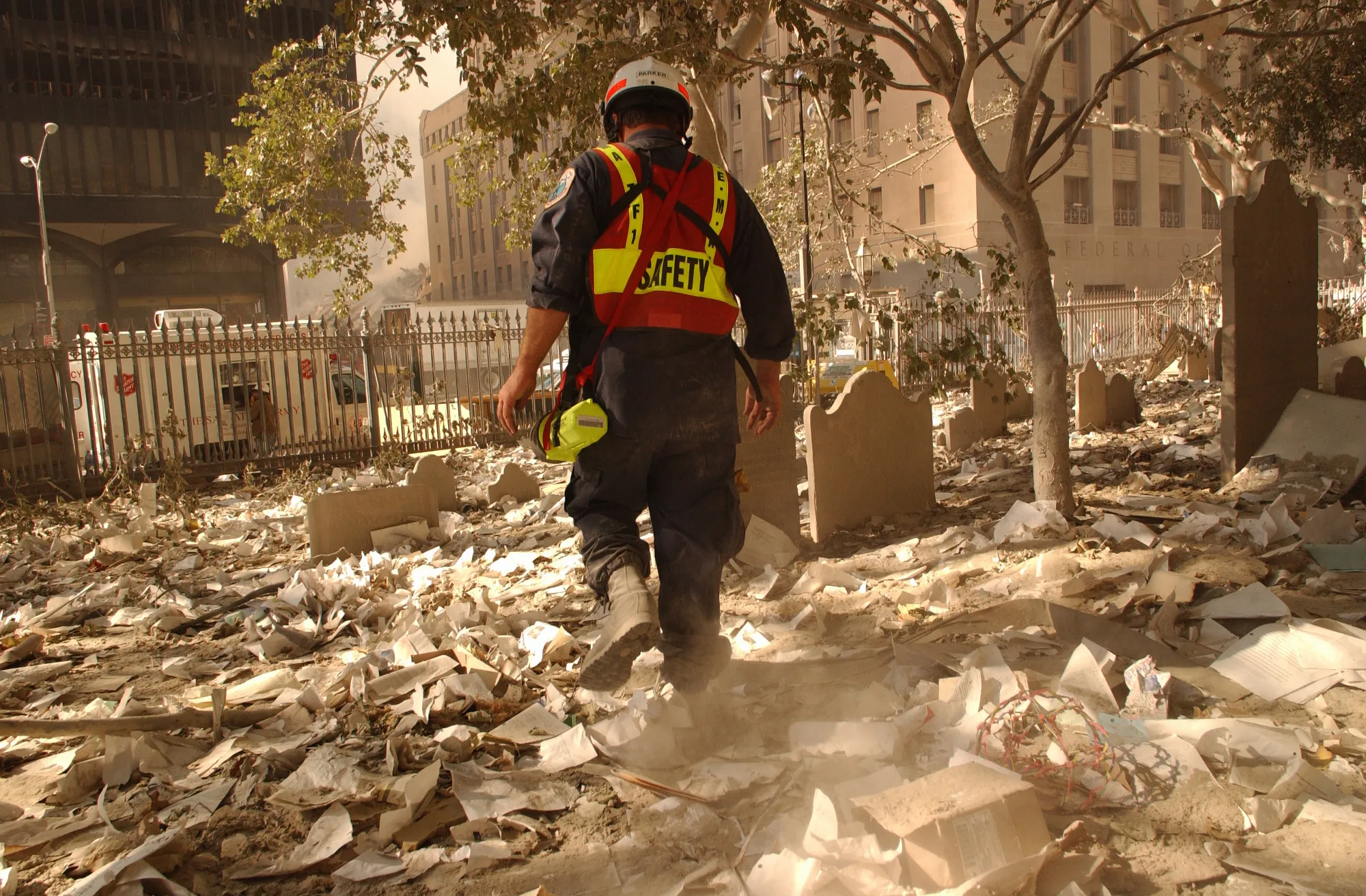 A man in a hardhat and orange vest labeled 'safety' walks through a NYC cemetery covered in dust and debris after 9/11