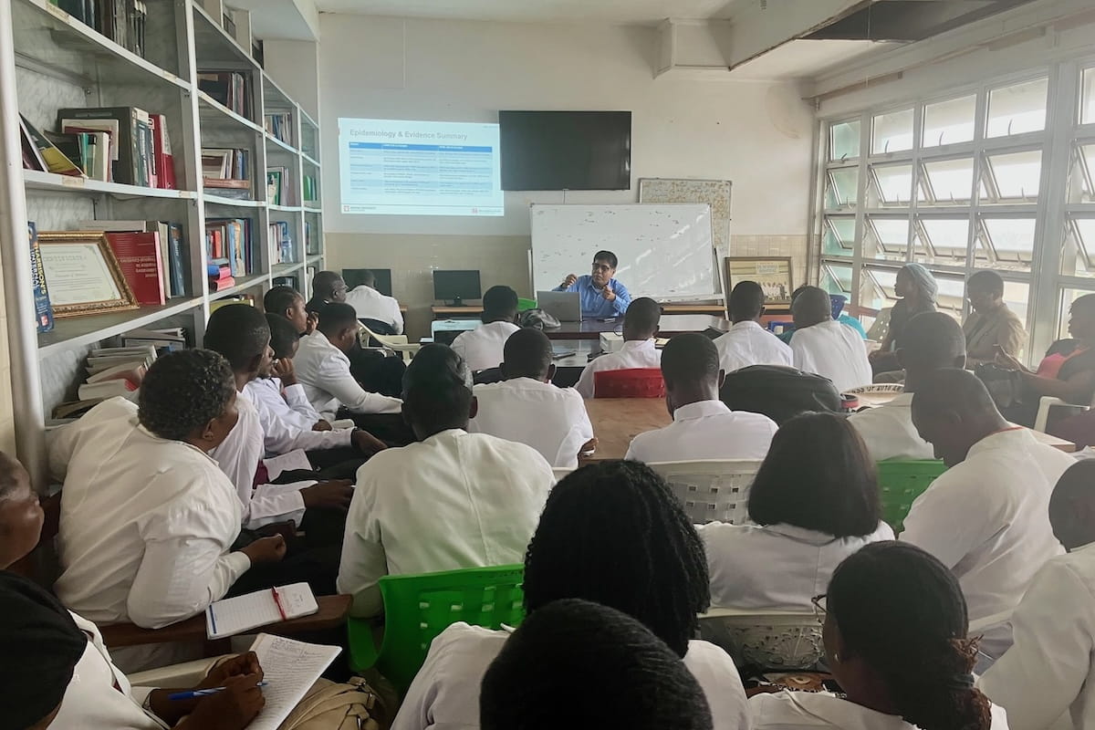 Adnan Bhutta sits in front of a screen and whiteboard lecturing a classroom full of Liberian health workers wearing white coats.