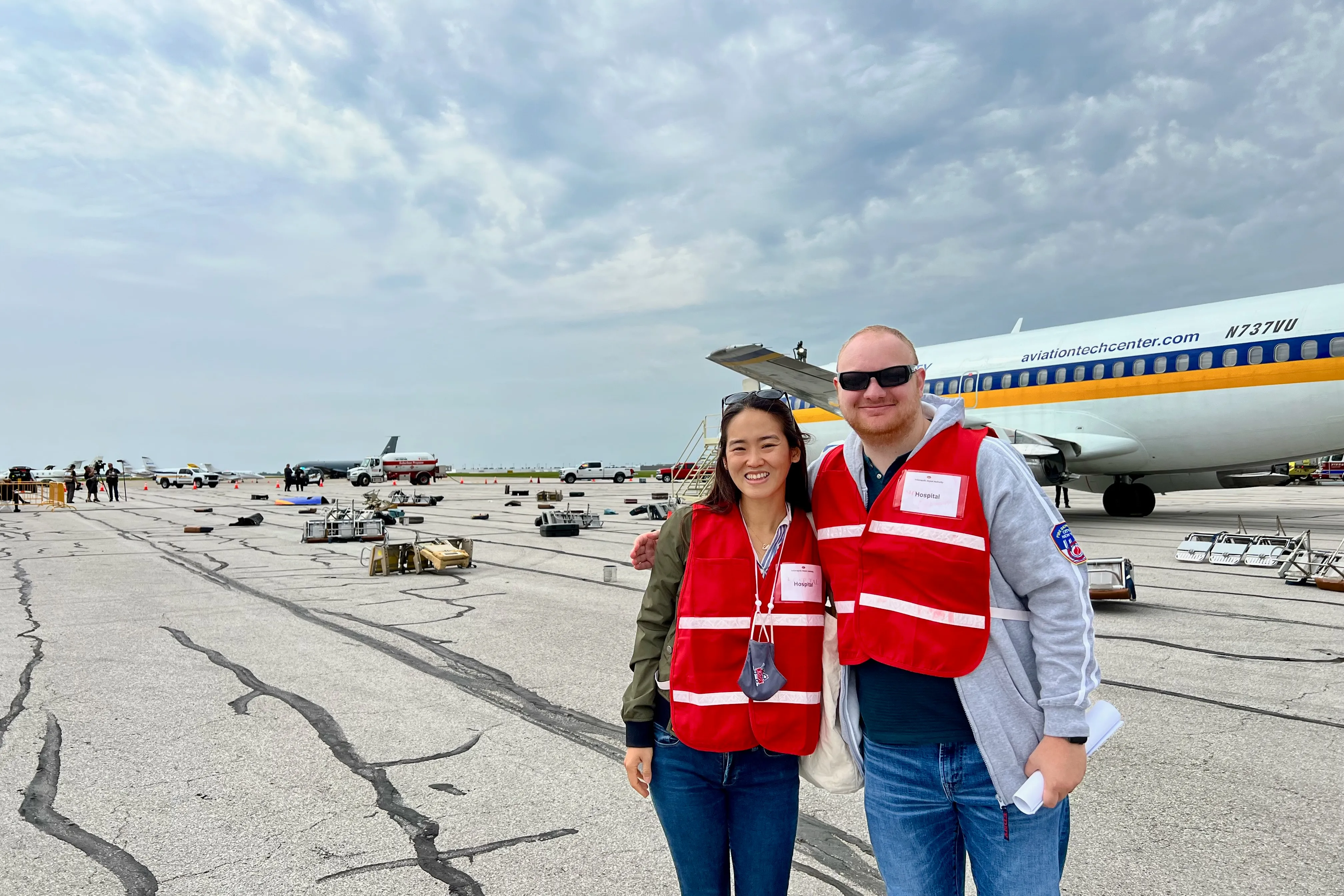 Two disaster medicine fellows in red vests, a woman and a man, stand in front of an airplane on the runway with ejected seats and baggage strewn about behind them..