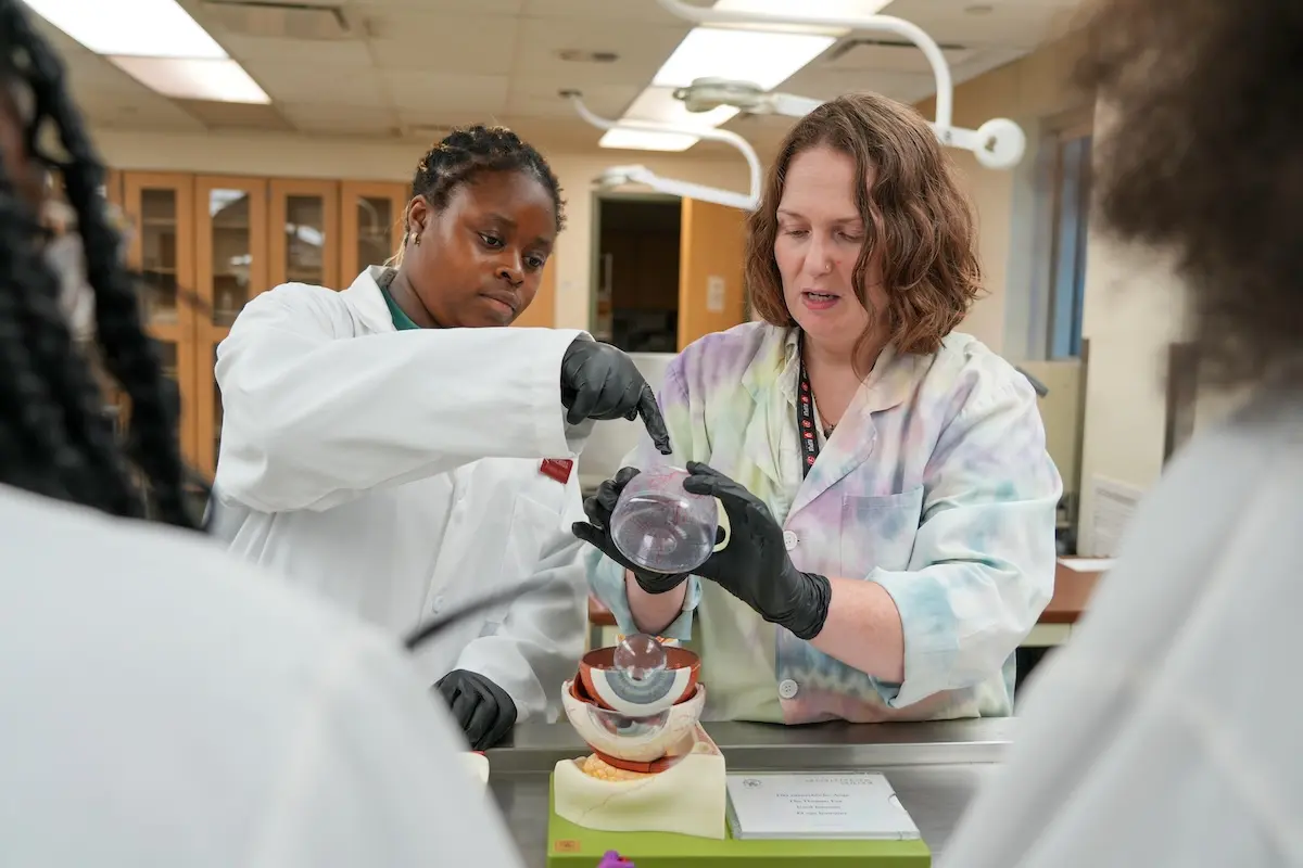 Liz Agosto, wearing a tie-dyed lab coat, helps a Black female high school student examine parts of the human eye using a 3D-printed model.