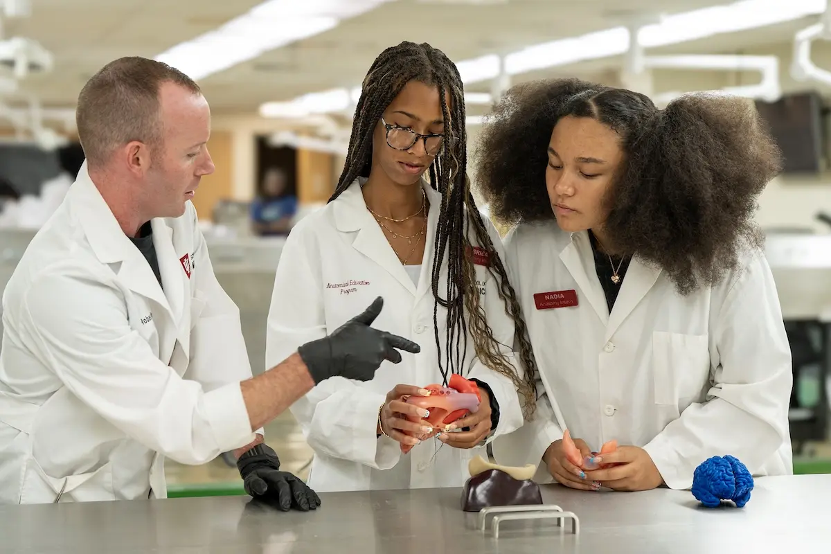 A male PhD student gestures at a heart model being inspected by two female high school students in the gross anatomy lab. All are wearing lab coats.