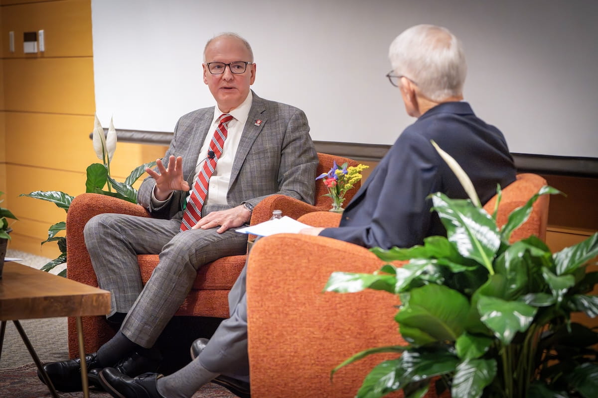 Jay Hess and Stephen Bogdewic sit in orange armchairs and converse at the front of a lecture hall.