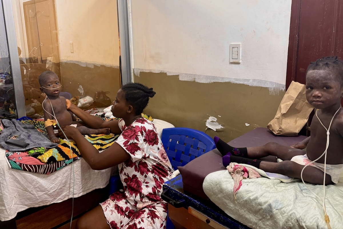 Two dark-skinned pediatric patients with nasal cannulas sit upright in hospital beds as one mother tends to her child. 