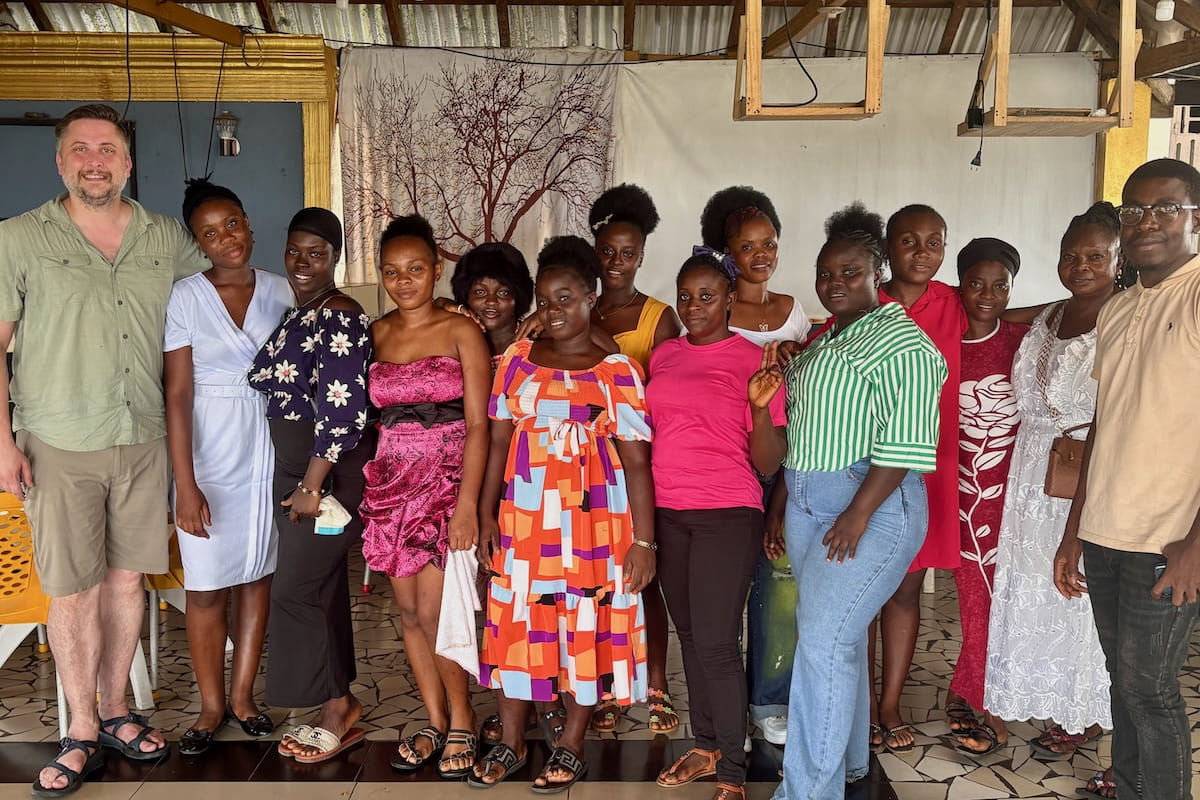 Mike Davis stands with 12 Liberian women in dress clothing and one Liberian man in a building with a tin roof.