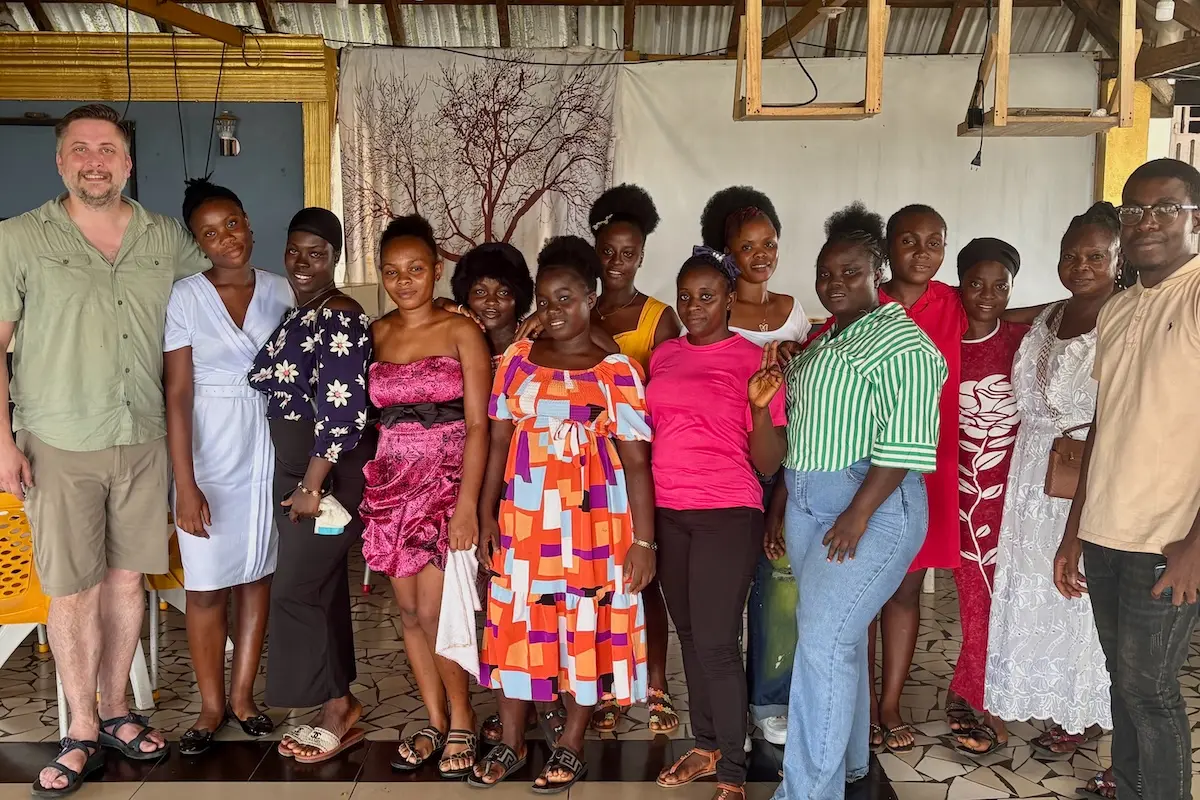 Mike Davis stands with 12 Liberian women in dress clothing and one Liberian man in a building with a tin roof.