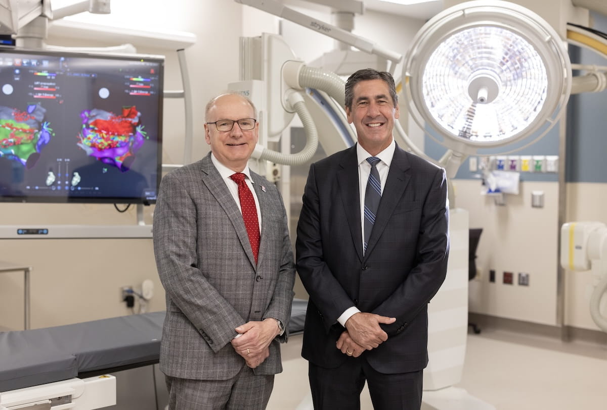 Jay Hess and Dennis Murphy stand together wearing suits in a clinical setting with large monitor and operating light