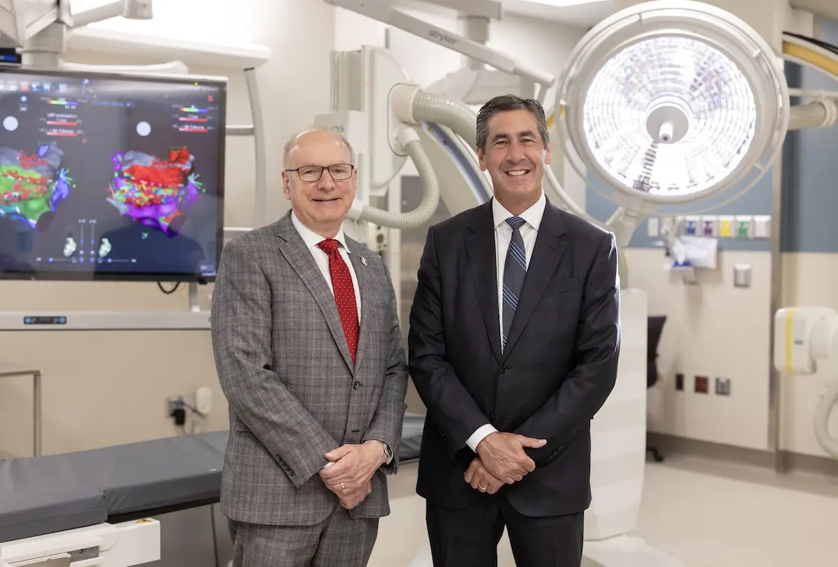 Jay Hess and Dennis Murphy stand together wearing suits in a clinical setting with large monitor and operating light
