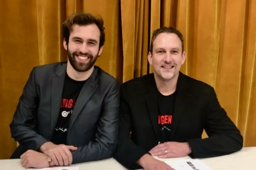 Jacob and Jeff Foy sit side-by-side at a table with a gold curtain behind them. They are both wearing Emergency the Musical T-shirts and dark colored blazers.