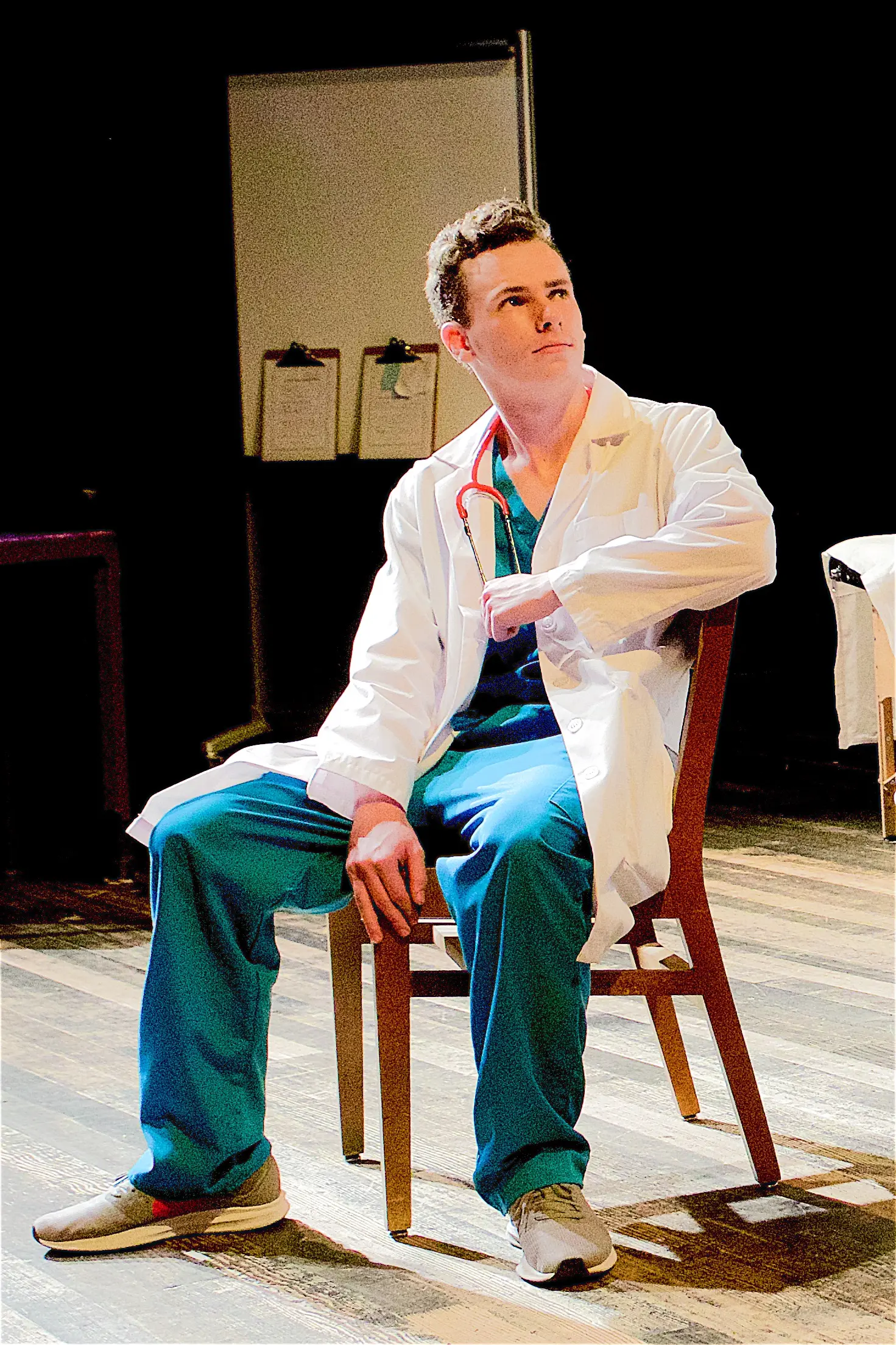 An actor portraying a young doctor sits in a chair in the spotlight on stage amid a set that's designed to look like a hospital.