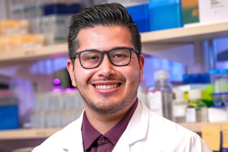 A man with dark hair and glasses smiles inside of a research lab. 