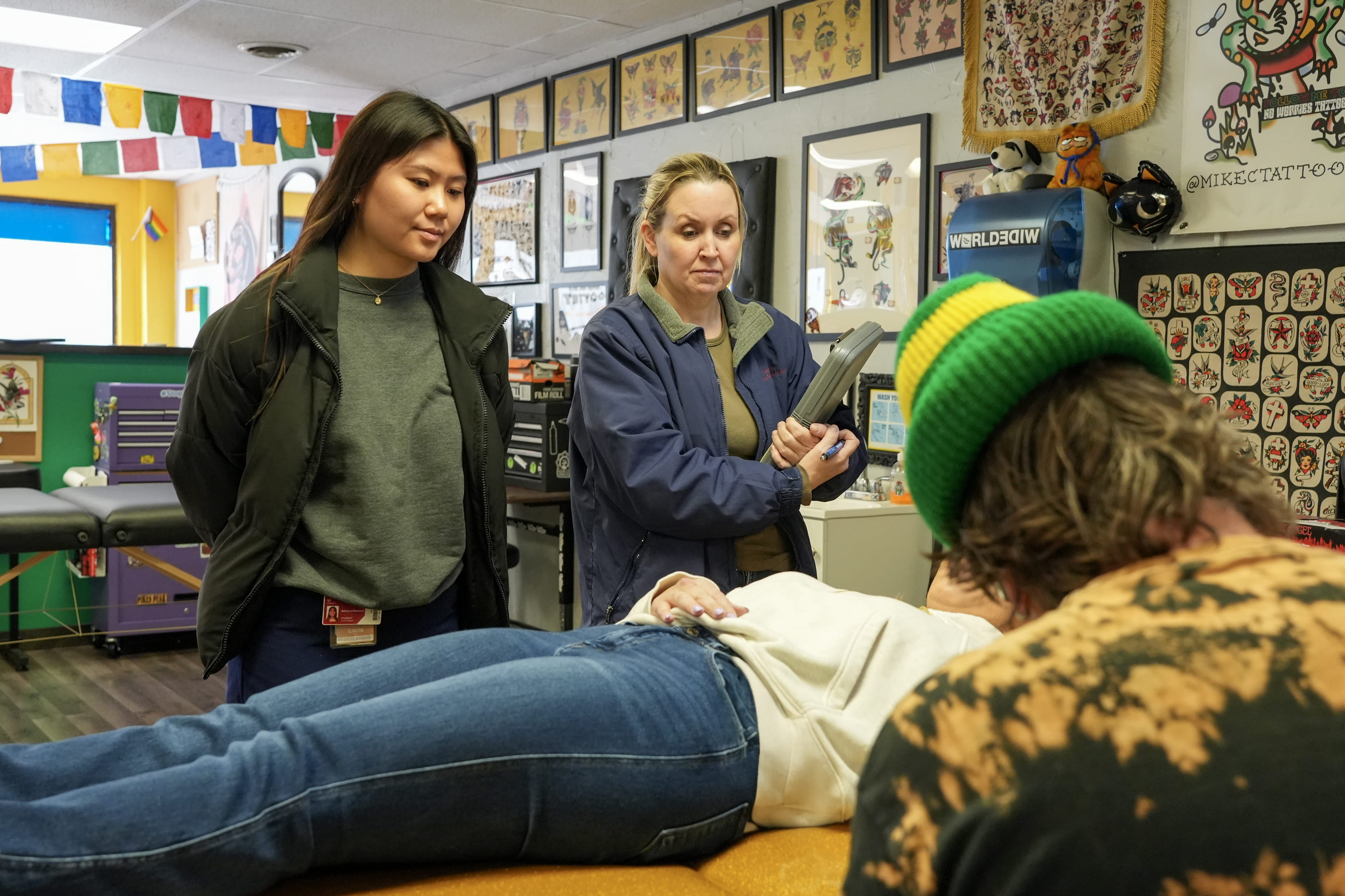 Lucia Li stand next to a health department worker and observes as a tattoo shop worker prepares to give a tattoo.