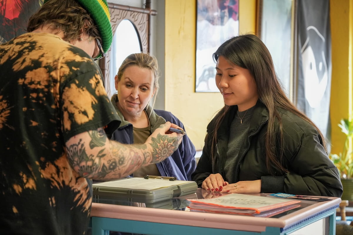 Lucia Li stands with a health department worker on opposite side of counter from a man with tattoos who is signing paperwork.