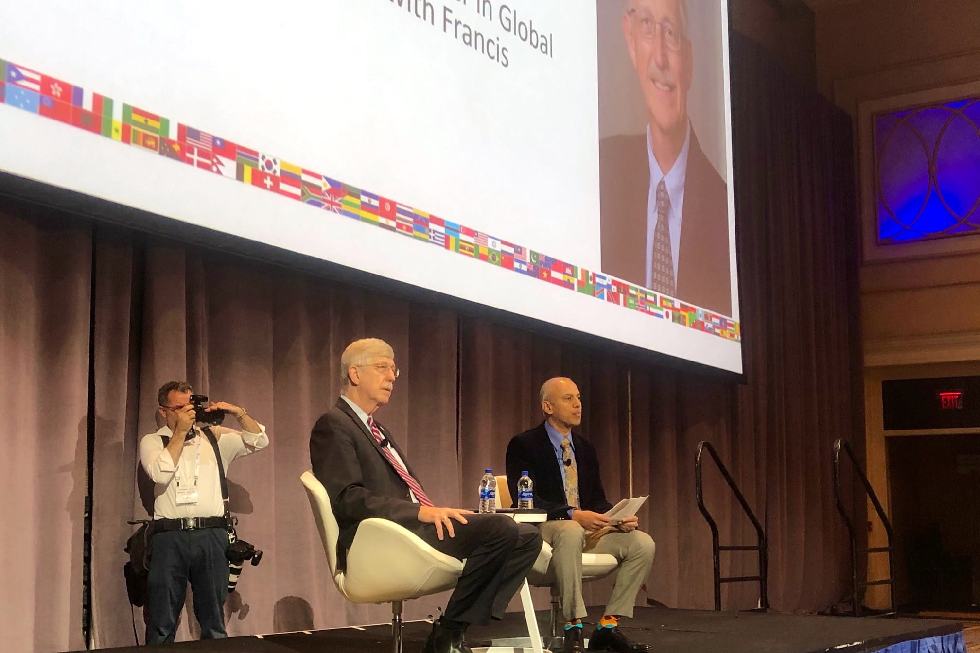 Francis Collins and Chandy John sit in chairs on stage as a photographer takes photos behind them. Francis Collins' photo appears on a big screen behind them.