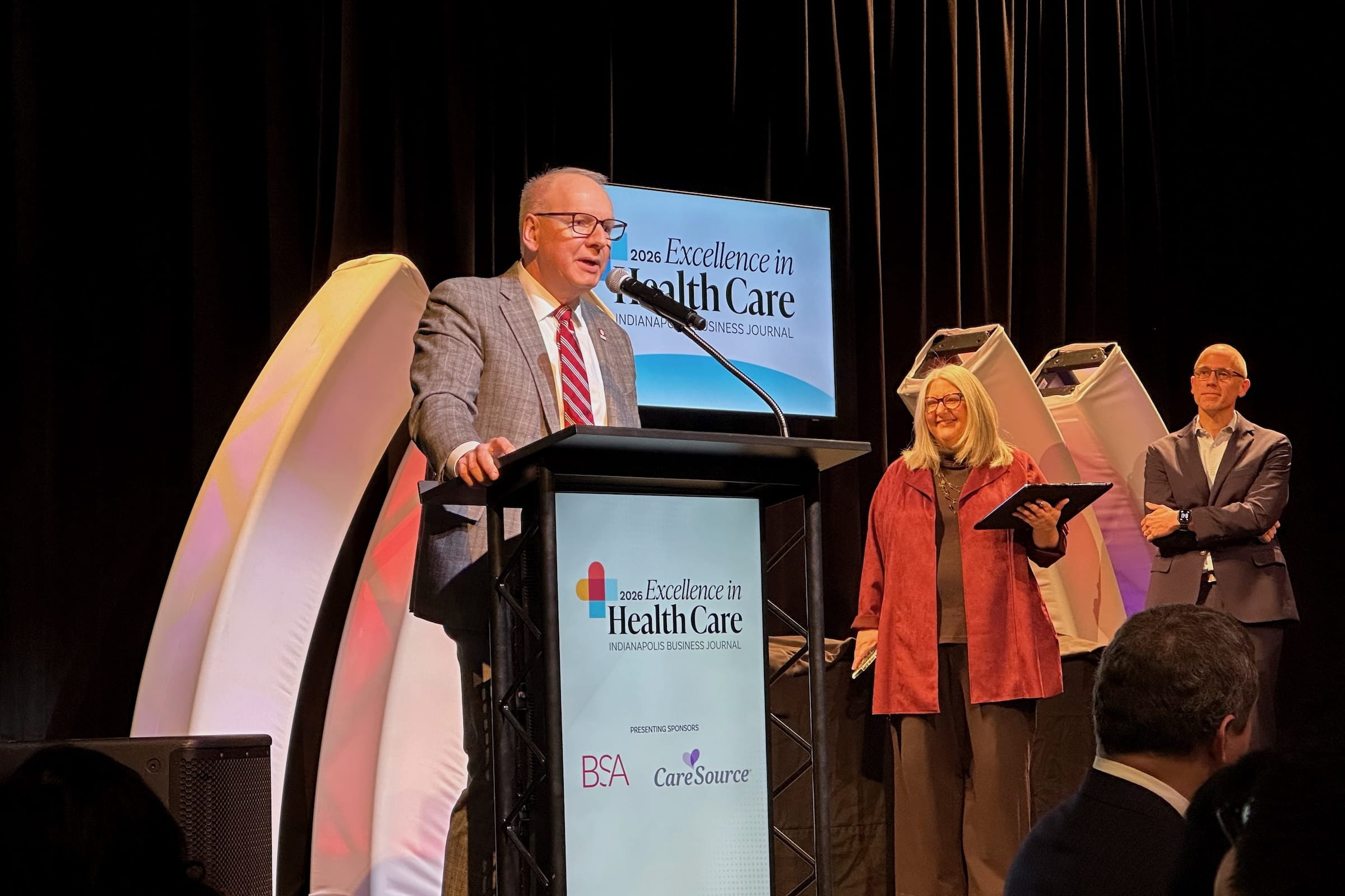 Dean Jay Hess speaks at podium with Excellence in Health Care logo as others watch