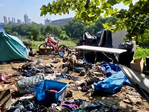 Tents and scattered personal belongings sit under the shade of a tree with the Indianapolis skyline in the background.