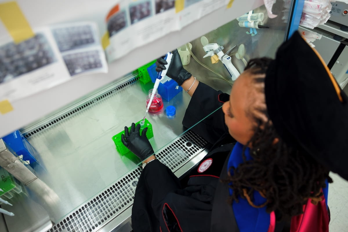 Ivana Daniels, in her graduation regalia, works with a pipet in the lab