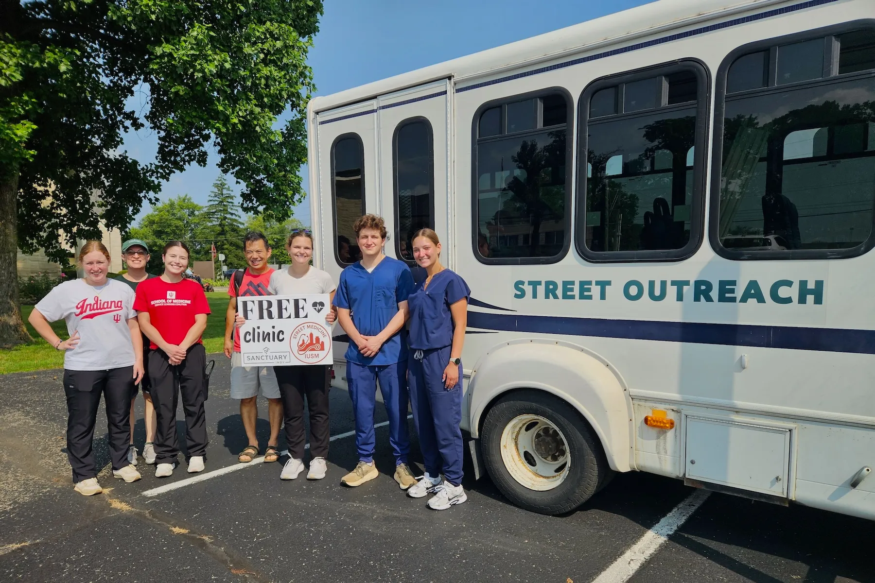 Ira Means and six MD students stand in front of a white bus labeled Street Outreach, holding a sign for Street Medicine at IU School of Medicine